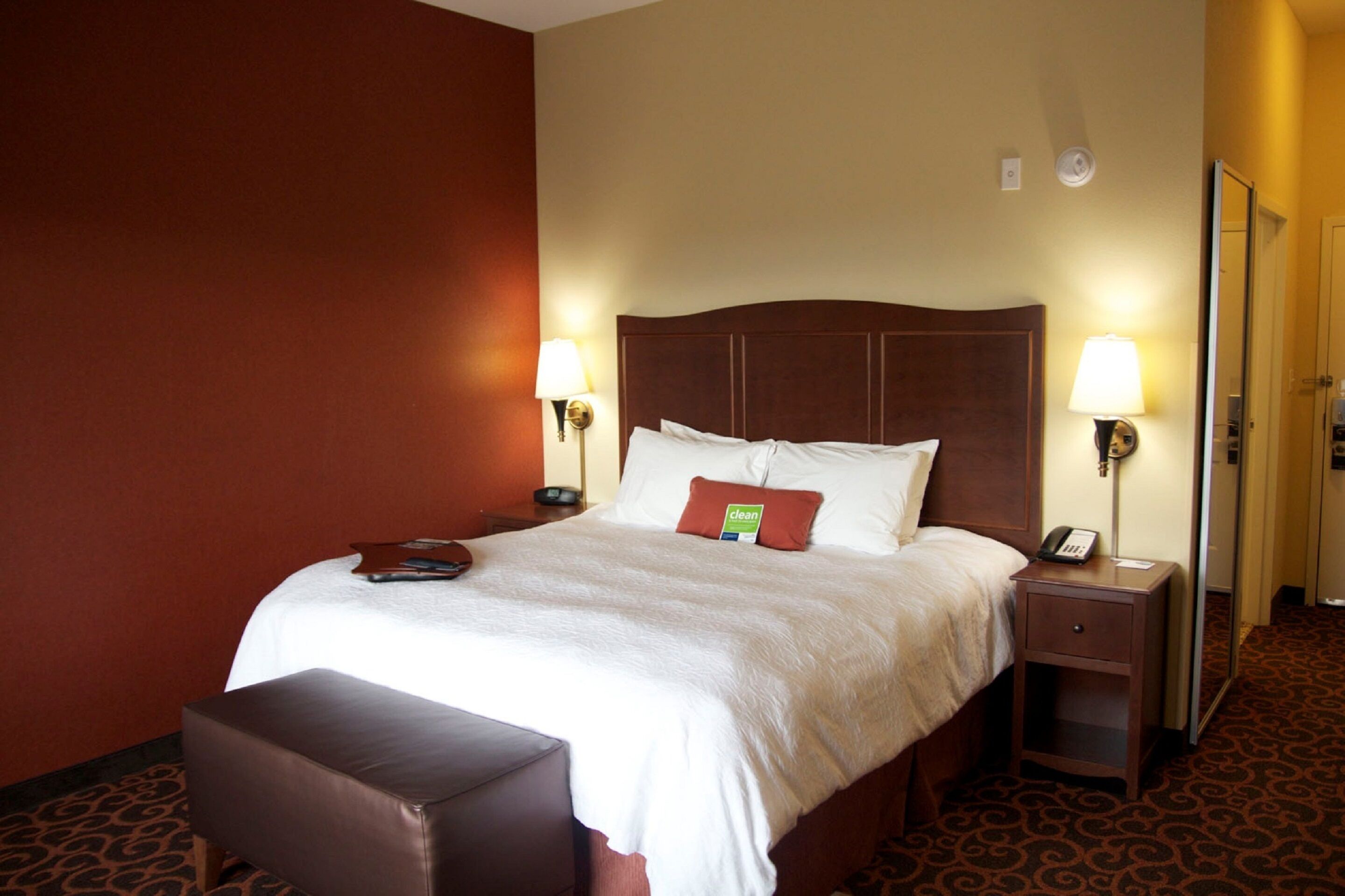 A hotel room with a large bed, two bedside tables with lamps, a small desk, and a brown leather bench.