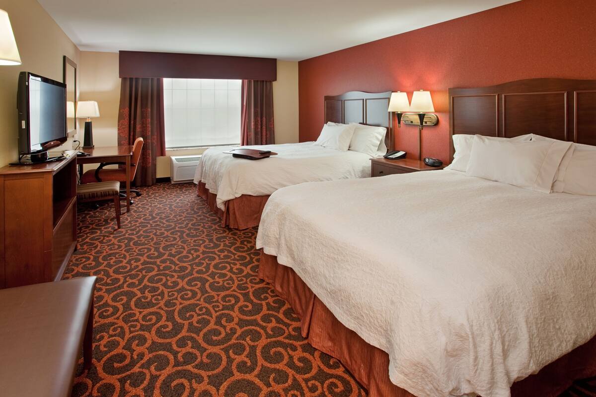 A hotel room with two beds, a desk with a computer, a TV, and a window with curtains.