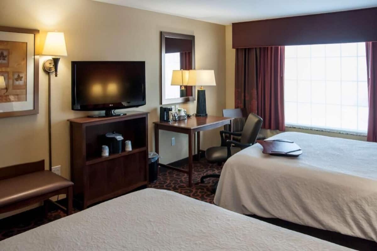 A hotel room with two beds, a television, a desk with a lamp, a chair, and a window with curtains.