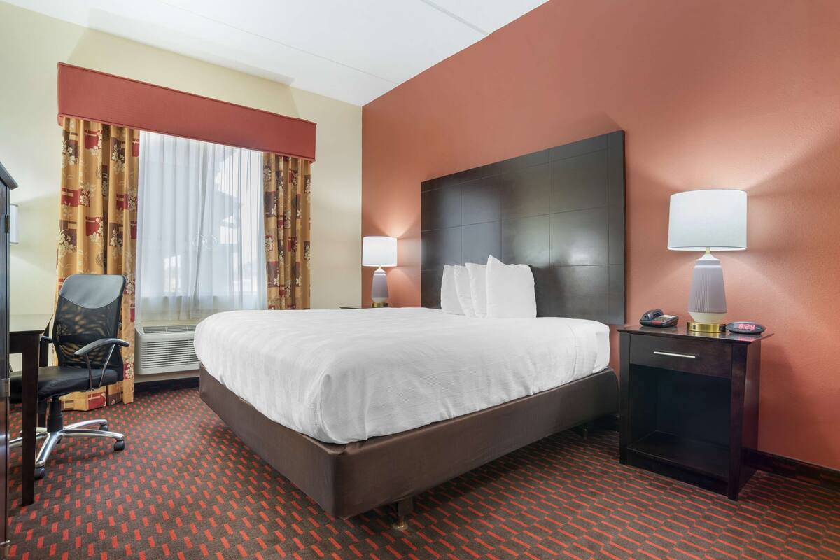 Ein Hotelzimmer mit einem großen Bett, einem Schreibtisch, einem Stuhl, zwei Nachttischlampen und einem Nachttisch mit Telefon und Wecker.