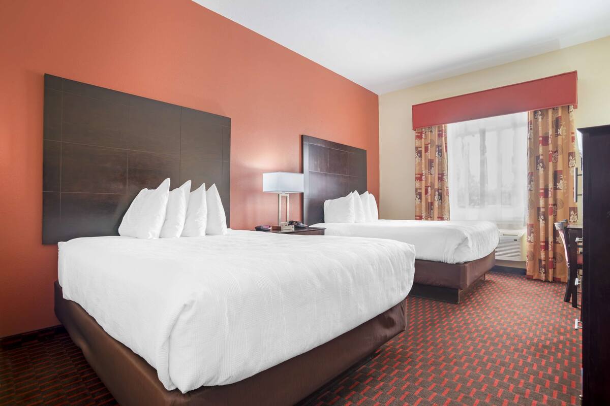 Ein Hotelzimmer mit zwei Betten, einem großen Kopfteil, einem Fenster mit Vorhängen und einem roten Teppich mit Muster.