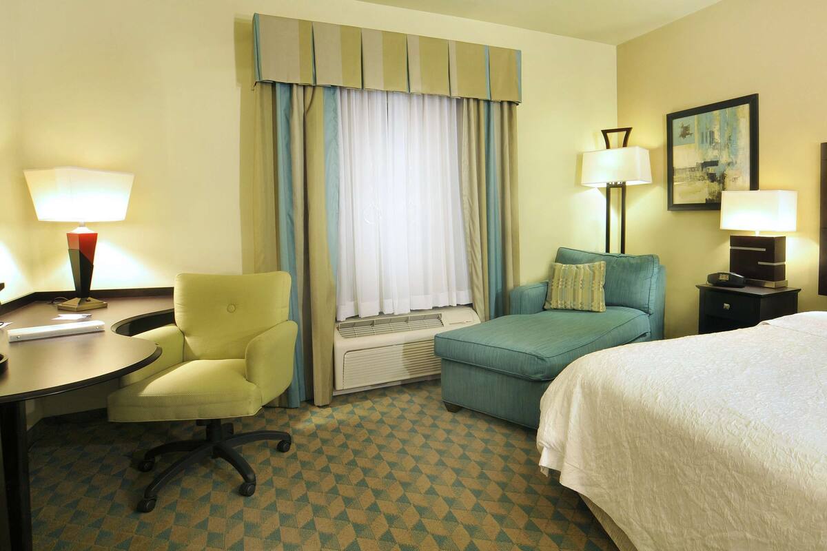A hotel room with a bed, a desk with a lamp, a chair, a sofa, and a window with curtains.