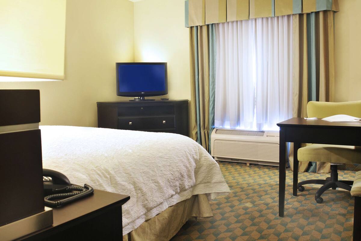 A hotel room with a bed, a desk with a chair, a television, a telephone, and a window with curtains.