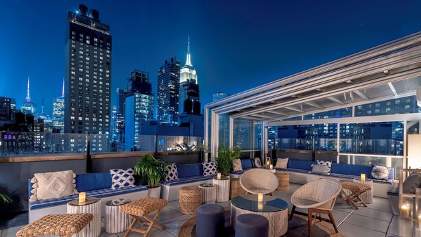 A rooftop terrace with a city skyline view, seating areas with cushions and tables, and a glass-enclosed section.