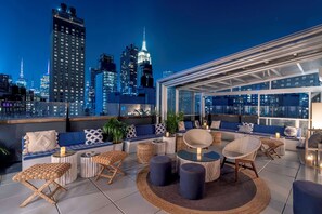 Una terraza en la azotea con vistas al horizonte urbano, zonas de estar con cojines y mesas, y una sección con cerramiento de cristal.