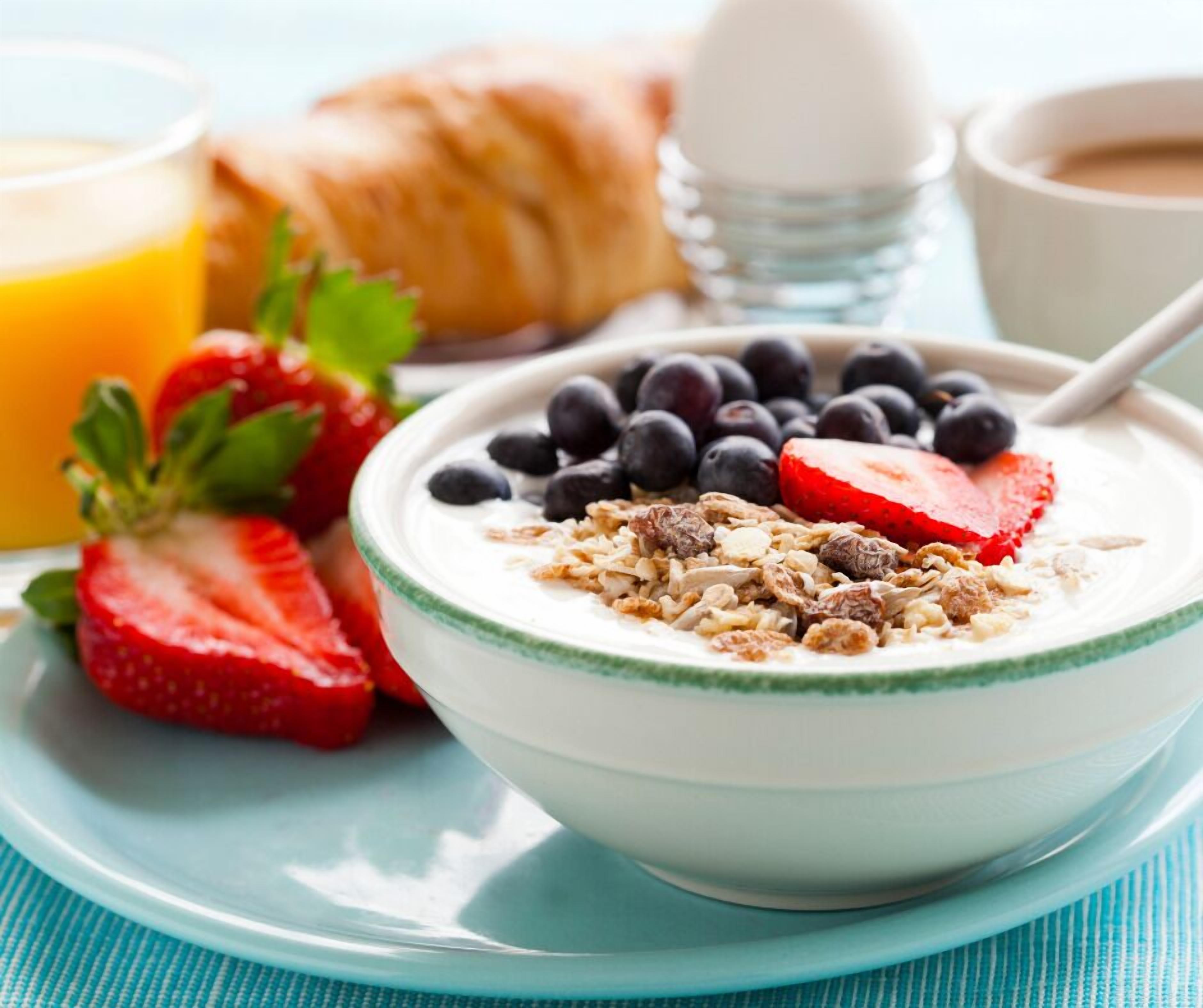 Een kom yoghurt met granola en bessen, een croissant, een glas sinaasappelsap en een kop koffie op een tafel.