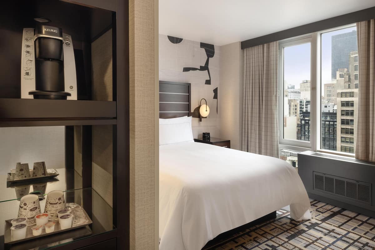 Habitación de hotel con cama, cafetera, mesita de noche, lámpara y ventana con vista a la ciudad.