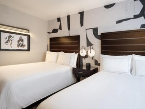 Two double beds with white linens are flanked by dark headboards, accompanied by a bedside table and a lamp.