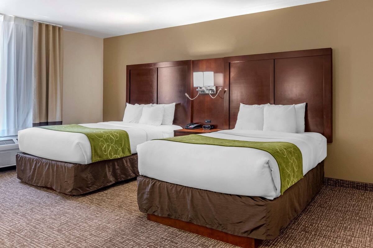 A hotel room with two beds, a wooden headboard, a bedside table with a phone, and a window with curtains.