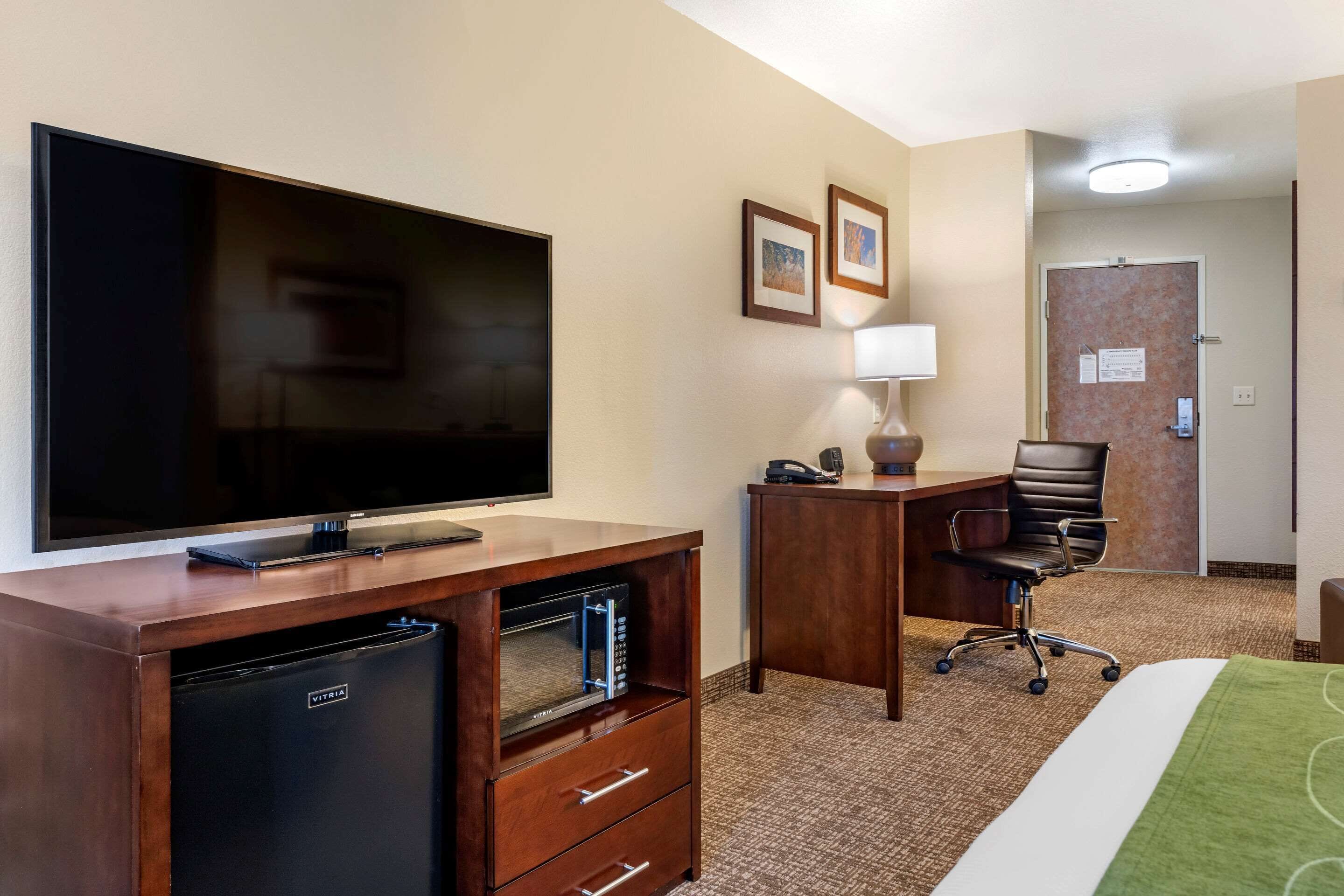 A hotel room with a flat-screen TV, a minibar, a desk with a lamp, a chair, and a bed with a green blanket.