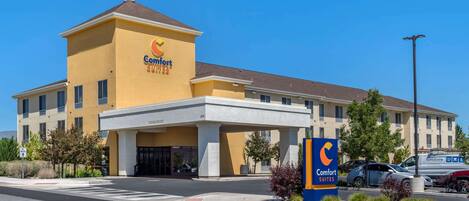 Un hotel Comfort Suites con fachada amarilla, un llamativo logotipo 'C' y un estacionamiento con varios automóviles.