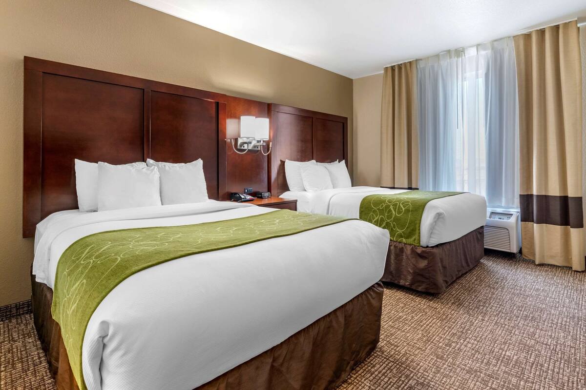 A hotel room with two beds, a wooden headboard, a bedside table with a lamp, and a window with curtains.