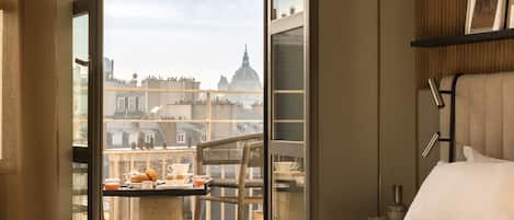 A hotel room with a large bed, a balcony with a table set for breakfast, and a city view.