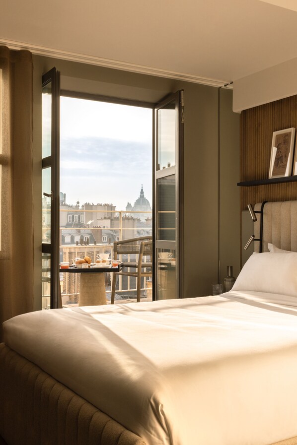 A hotel room with a large bed, a balcony with a table set for breakfast, and a city view. - Hotel Dame des Arts (Paris)