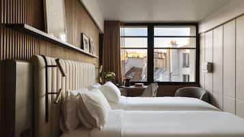 A hotel room with a large bed, a desk with a chair, a window with a view of buildings, and a wall-mounted shelf with decorative items.