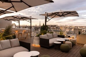 A rooftop terrace with a sofa, chairs, and tables under umbrellas, overlooking a cityscape with a prominent tower.