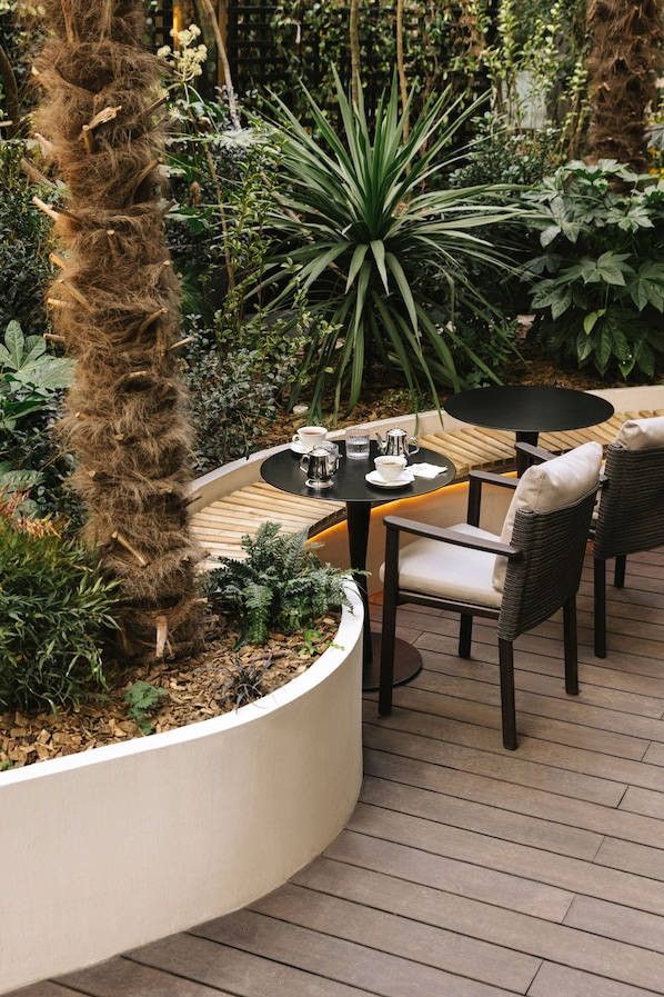A patio with a round table and two chairs, surrounded by lush greenery and a tall tree. - Hotel Dame des Arts (Paris)