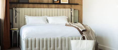 Una cama bien hecha con cabecera, una mesita de noche con lámpara y un bolso blanco grande en el suelo.
