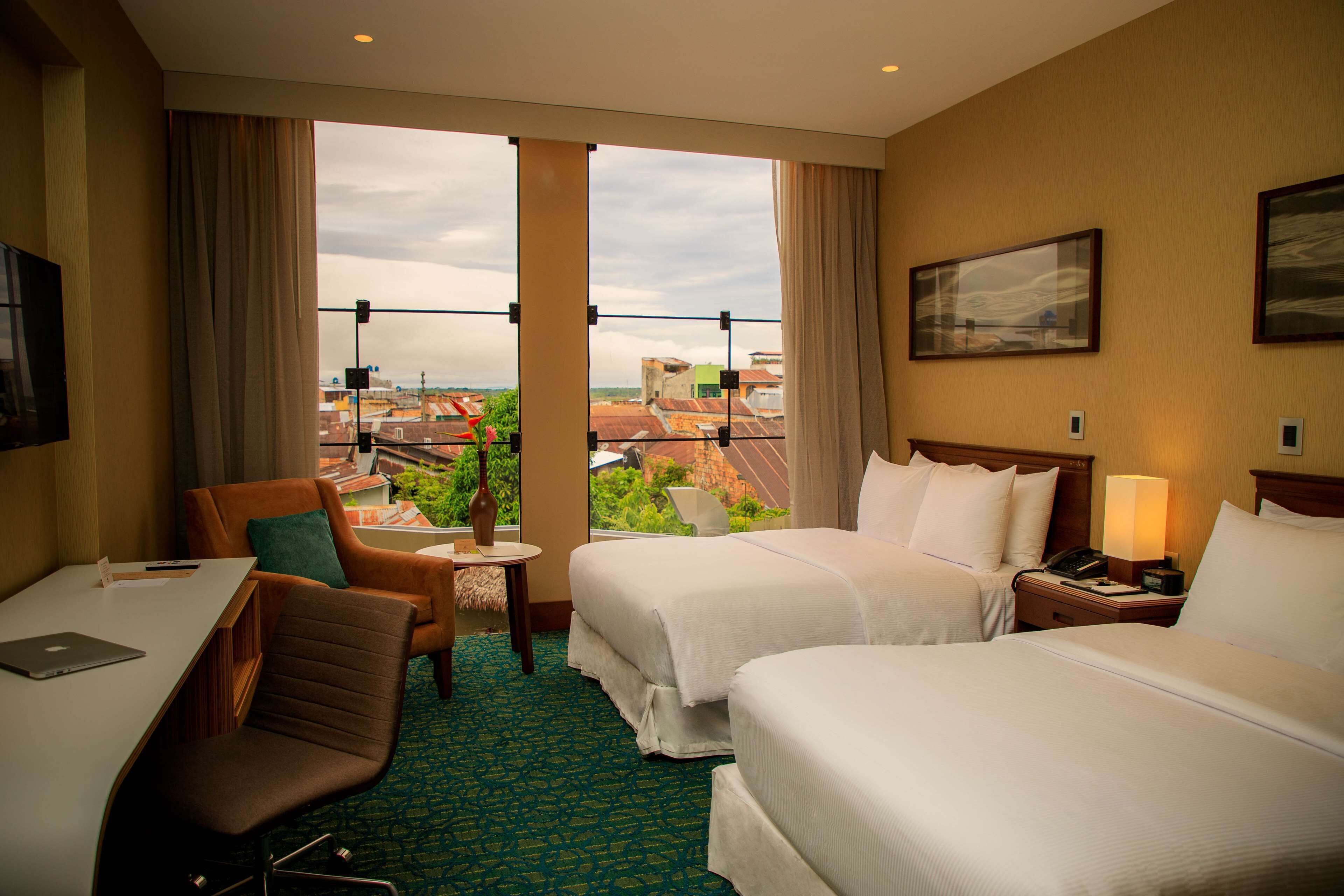 Habitación de hotel con dos camas, un escritorio con una computadora portátil, una silla, una mesita con un jarrón y vistas a edificios a través de grandes ventanales.