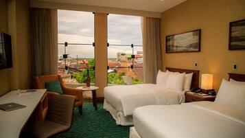 Habitación de hotel con dos camas, un escritorio con una computadora portátil, una silla, una mesita con un jarrón y vistas a edificios a través de grandes ventanales.