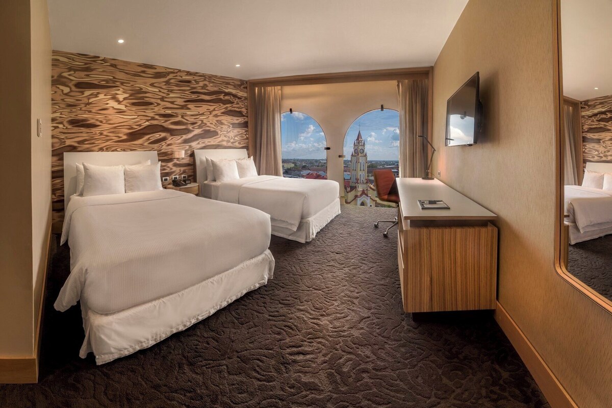 Quarto de hotel com duas camas, uma escrivaninha, uma cadeira e um espelho grande.