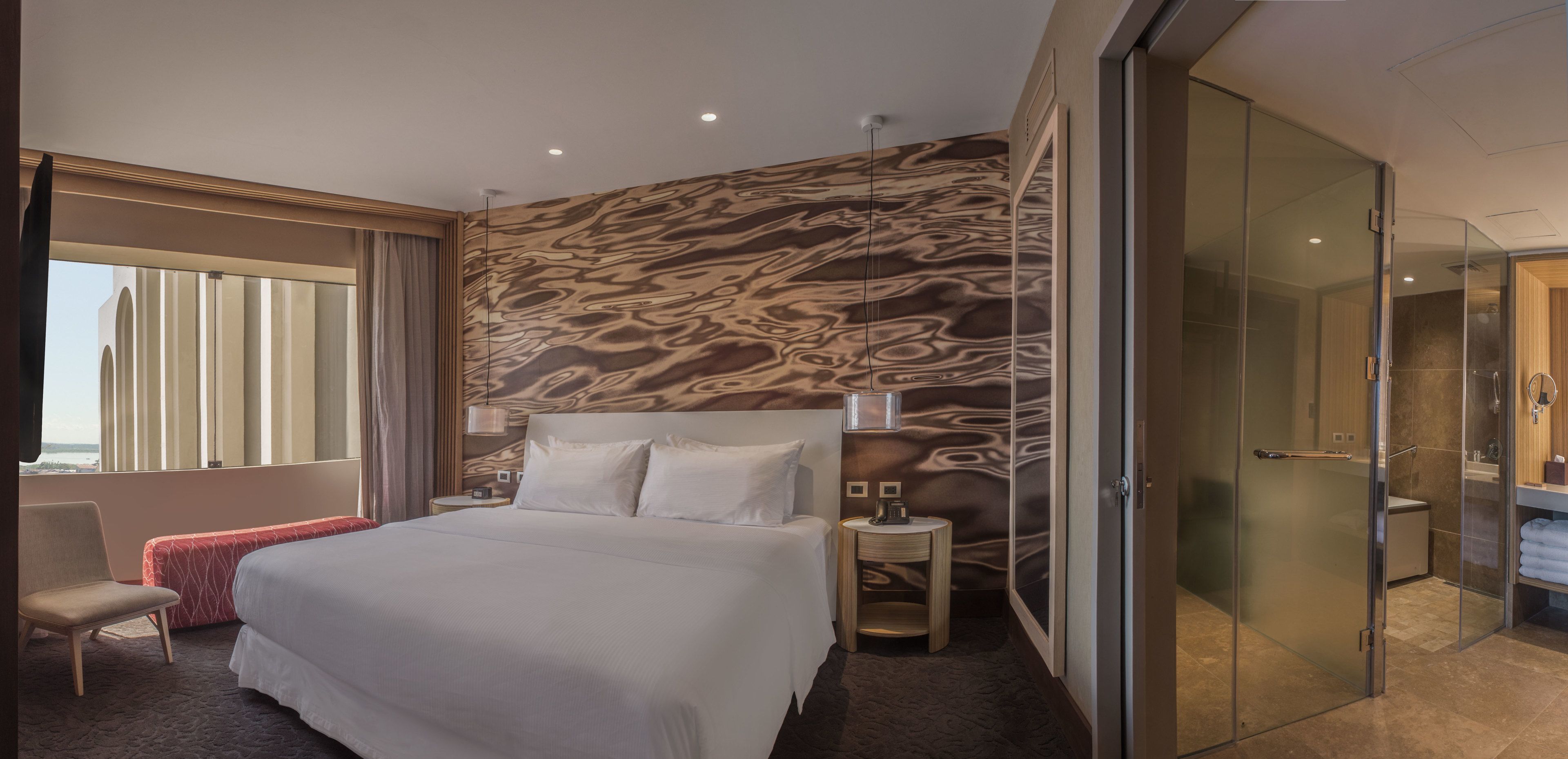 A hotel room with a large bed, a bedside table, a lamp, a red bench, a glass-enclosed shower, and a view of the sea.