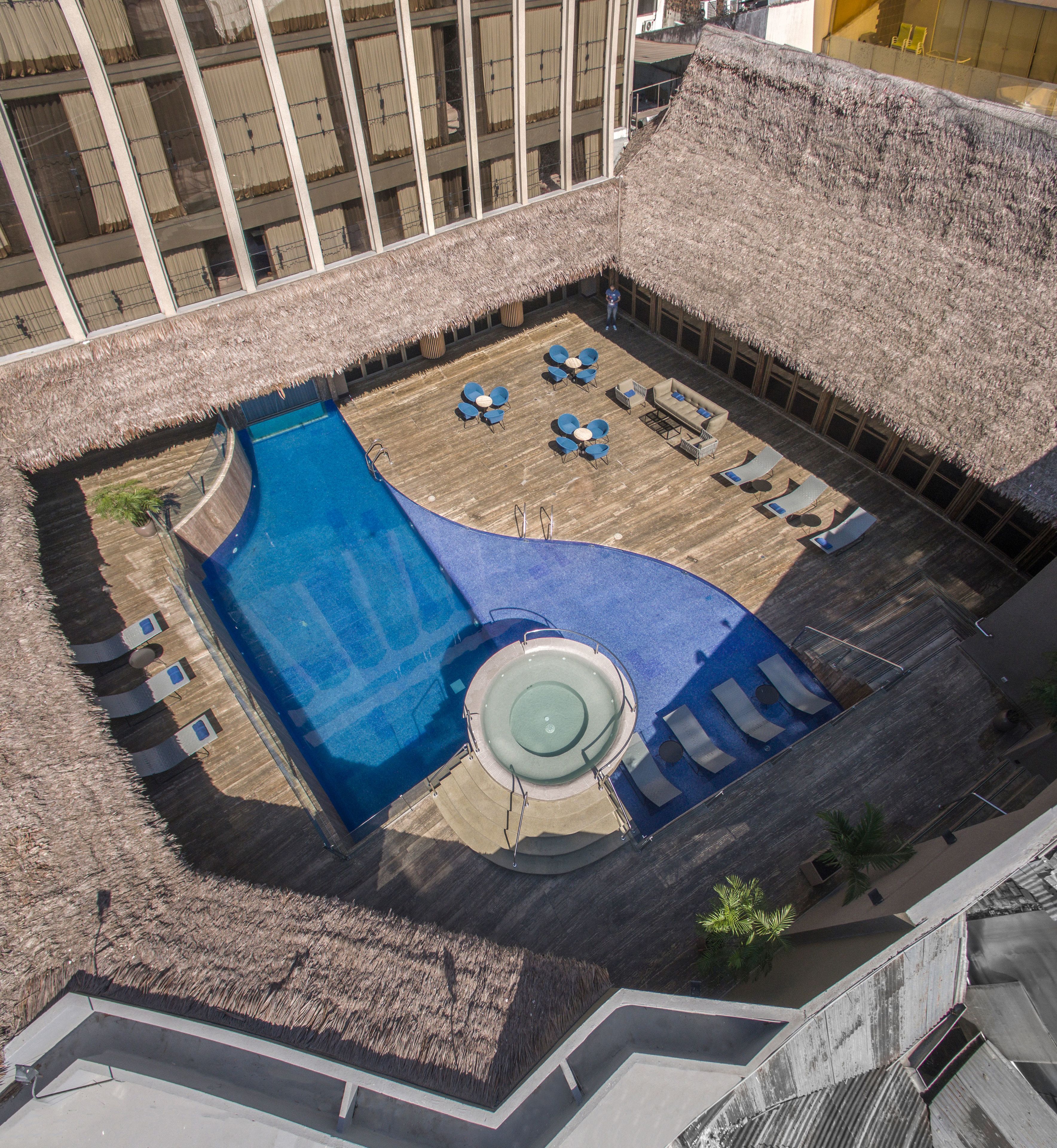An aerial view of a rooftop pool area with a circular hot tub, lounge chairs, and a thatched roof.