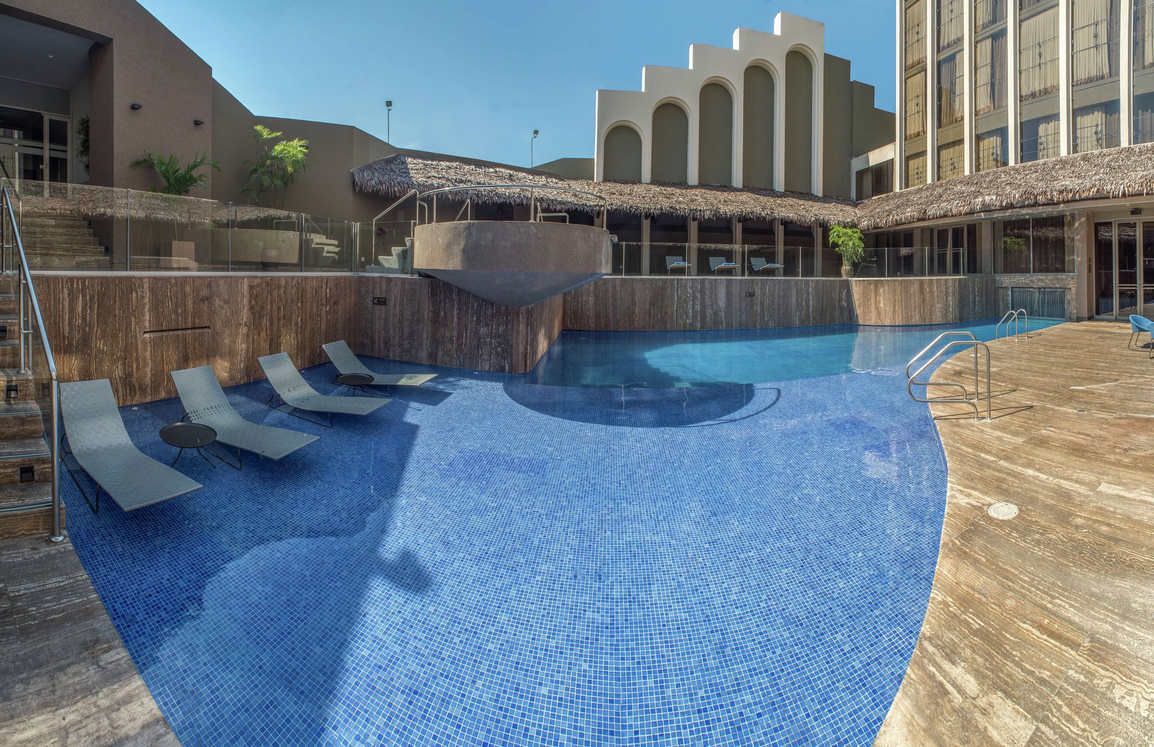 Una piscina con sillas de descanso, una fuente de agua y un edificio con ventanas de arco y techo de paja.