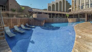 Una piscina con sillas de descanso, una fuente de agua y un edificio con ventanas de arco y techo de paja.