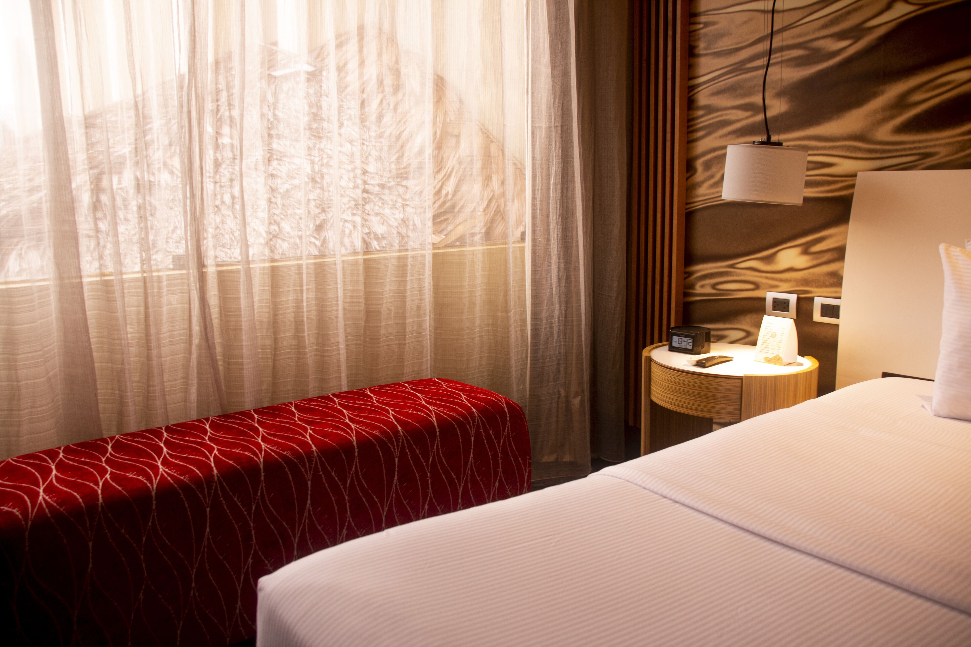 Quarto de hotel com uma cama, uma mesa de cabeceira com abajur, um banquinho estofado vermelho e cortinas leves.
