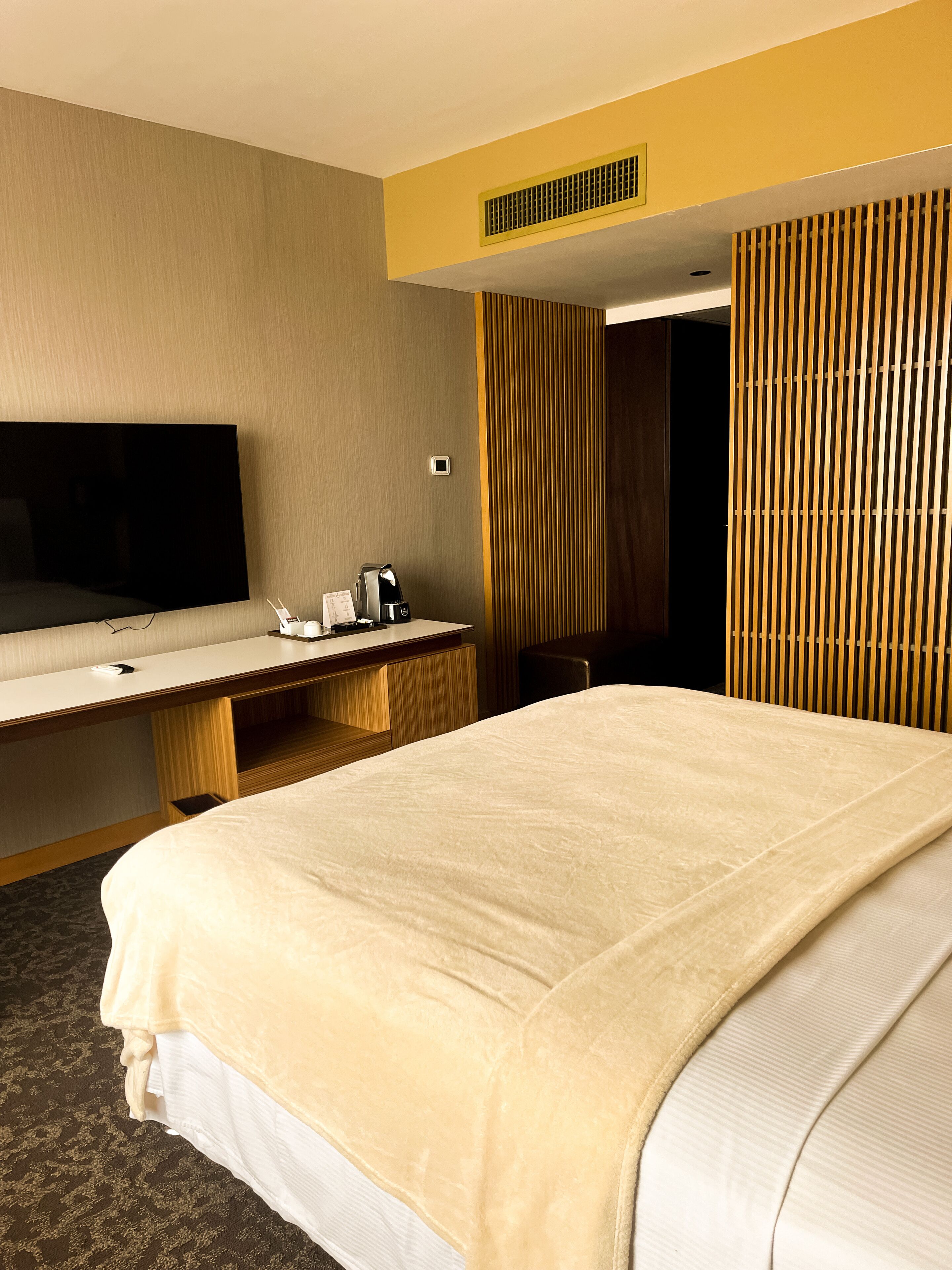 Habitación de hotel con una cama grande, un escritorio con televisión de pantalla plana, cafetera y un ventilador fijado a la pared.