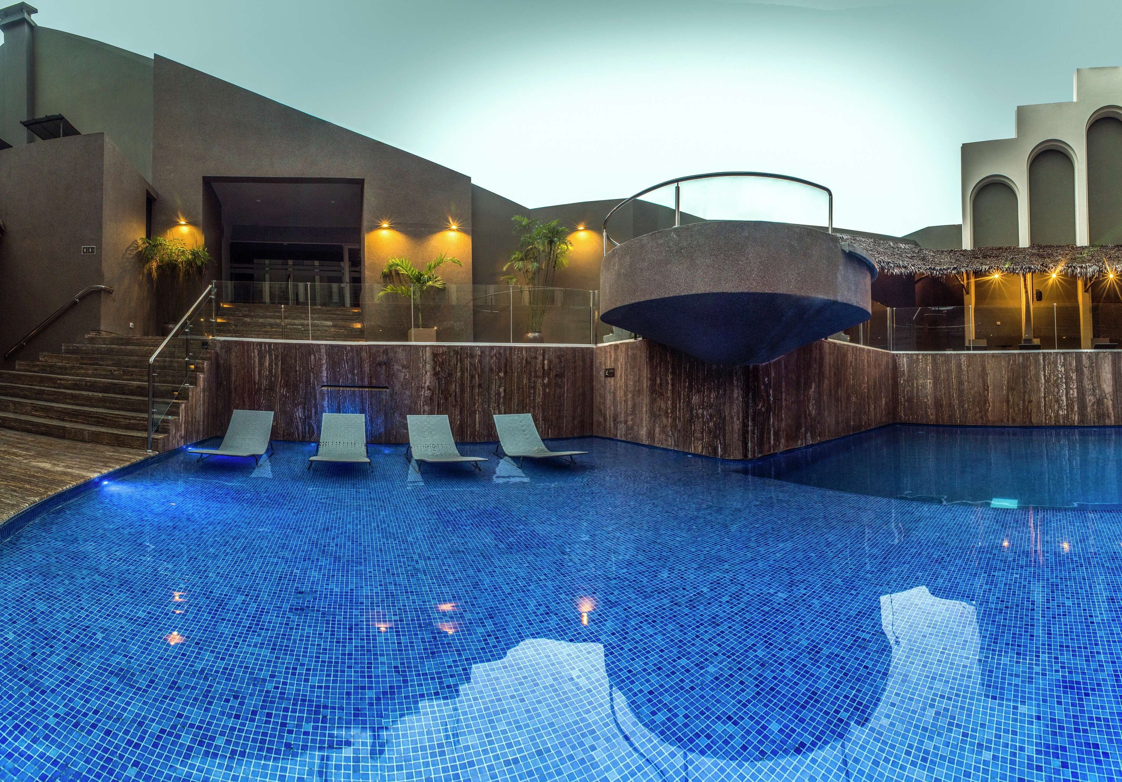 A modern indoor swimming pool with a wooden deck, lounge chairs, and a staircase leading to an upper level.