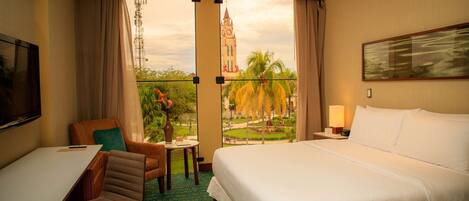 A hotel room with a large bed, a desk, a chair, and a view of a tower and palm trees.