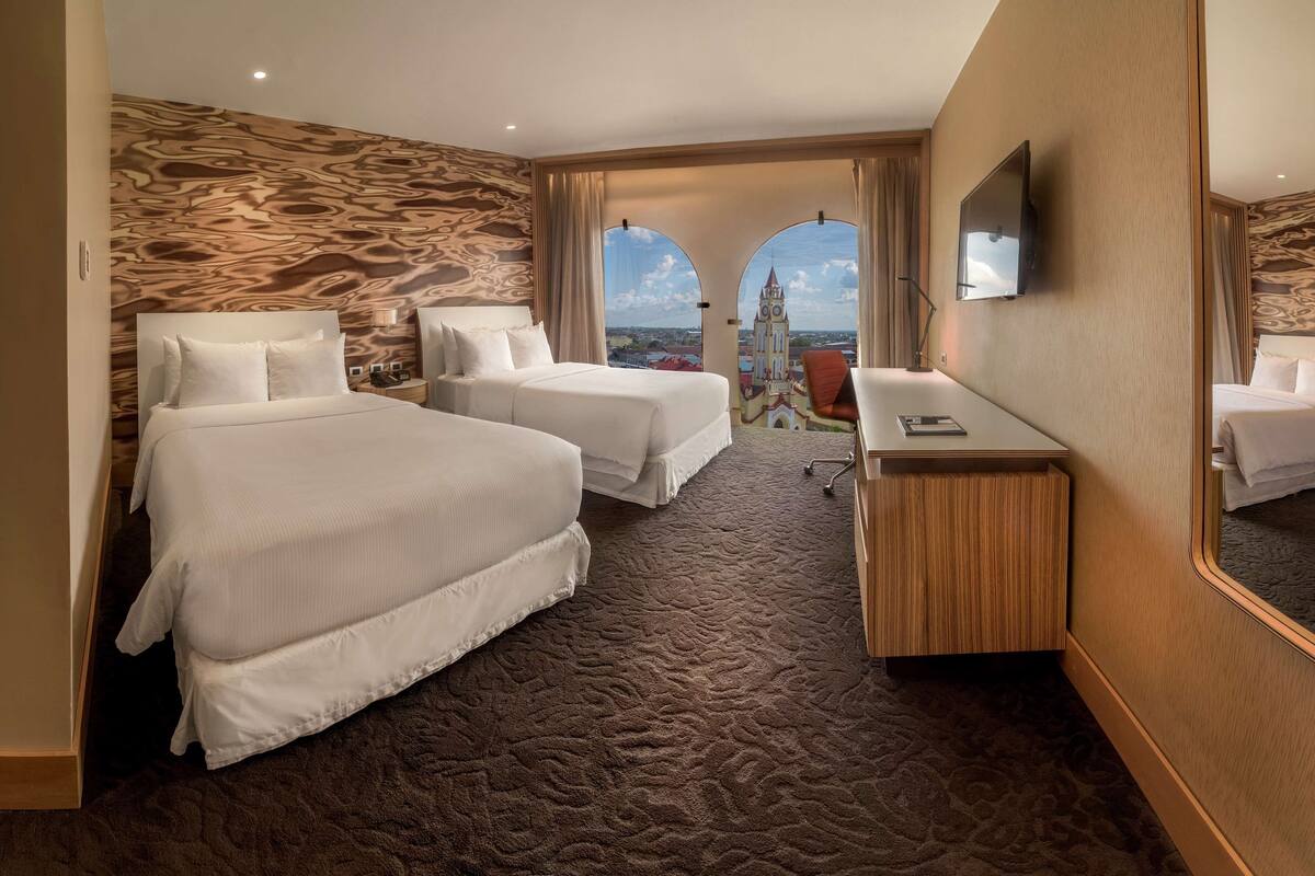 Ett hotellrum med två sängar, ett skrivbord, en TV och utsikt över en kyrka genom välvda fönster.
