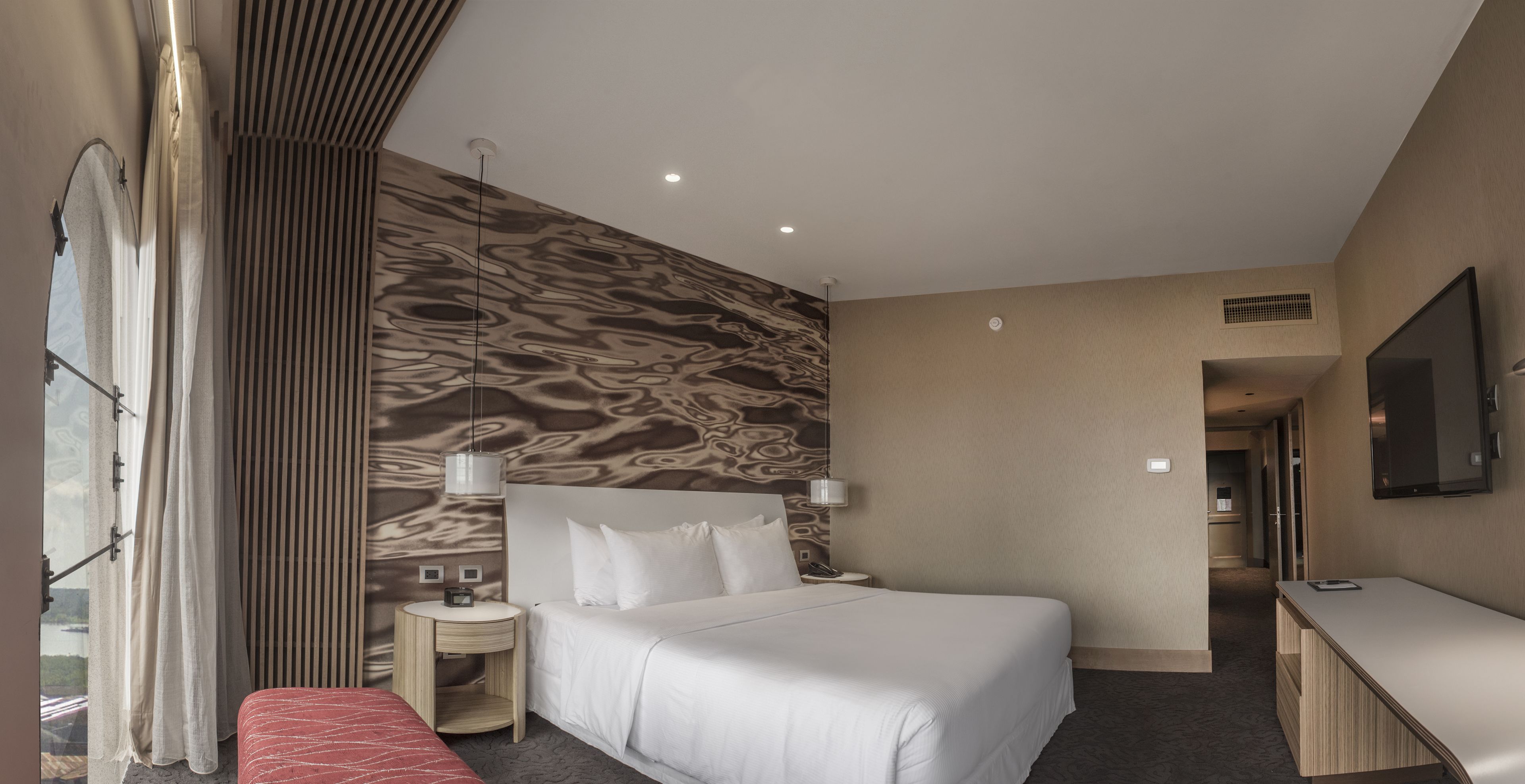 A hotel room with a large bed, a bedside table with a lamp, a TV, and a view of the outdoors.