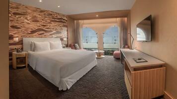 A modern hotel room with a large bed, a desk, and a view of the cityscape through large windows.