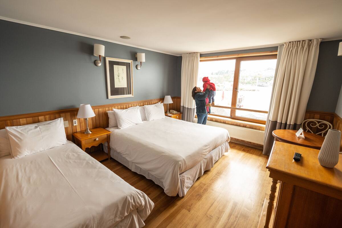 A hotel room with two beds, a wooden headboard, a nightstand with a lamp, a desk with a vase, and a window with curtains.