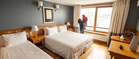 Una habitación de hotel con dos camas, un cabecero de madera, una mesita de noche con una lámpara, un escritorio con un florero y una ventana con cortinas.