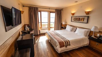 Ein Hotelzimmer mit einem großen Bett, Holzboden, einem Schreibtisch mit Stuhl, einem an der Wand montierten Fernseher und einem Balkon mit Vorhängen.