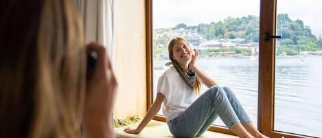 Una persona sentada junto a una ventana con vista a un cuerpo de agua y una ladera.