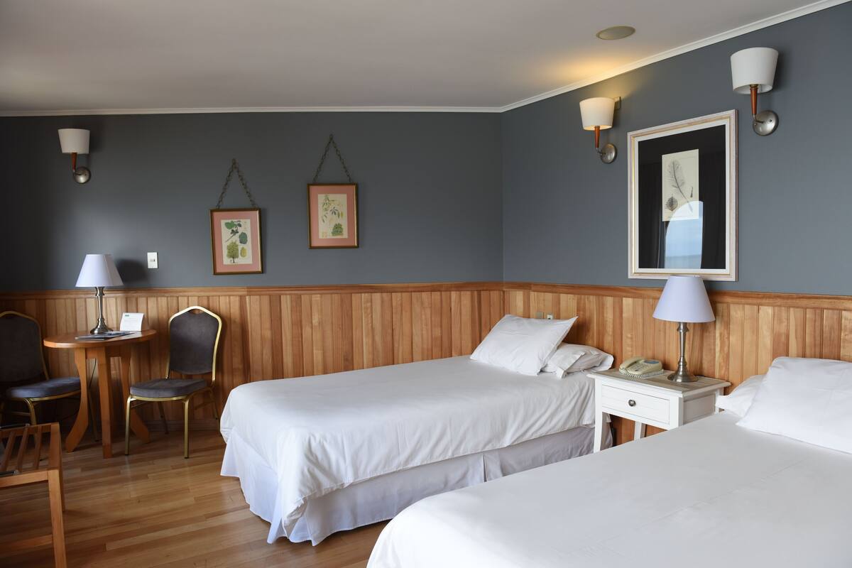 Une chambre d’hôtel avec deux lits, une tête de lit en bois, une petite table avec une lampe, une chaise et deux lampes fixées au mur.