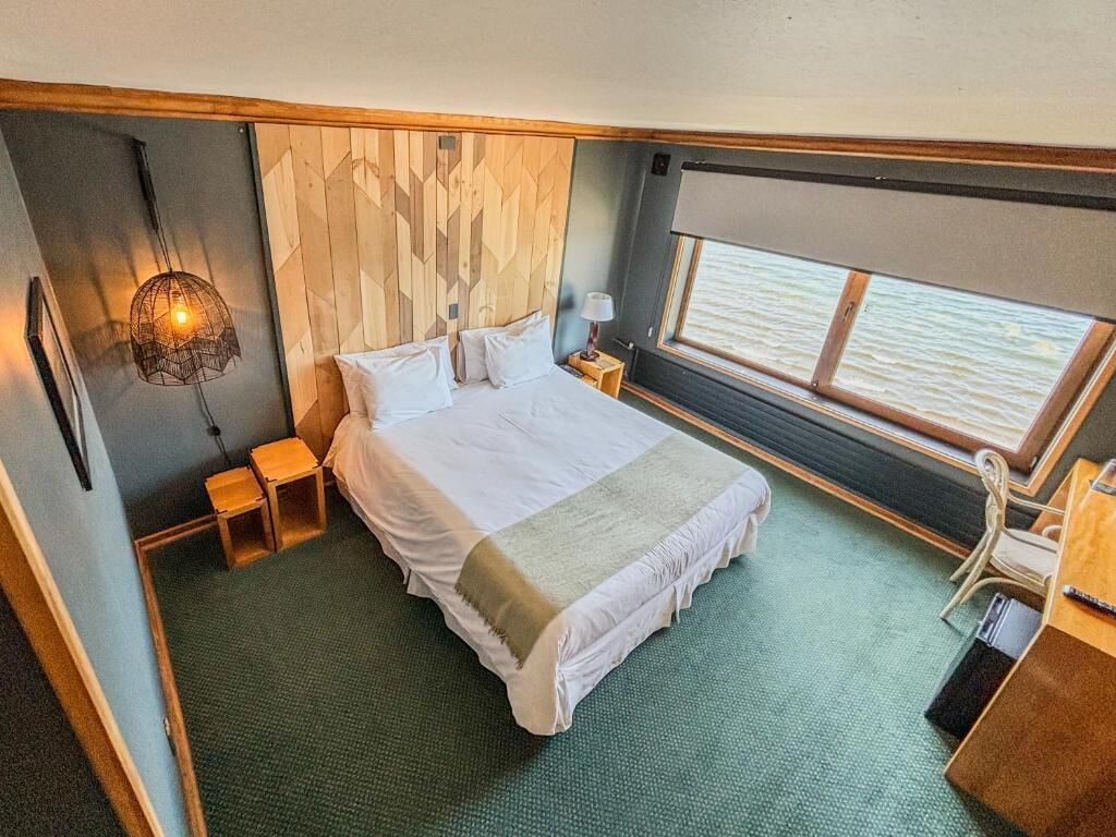 Un dormitorio con una cama, una mesita de noche, una lámpara, una pequeña mesa y una ventana con vista al agua.
