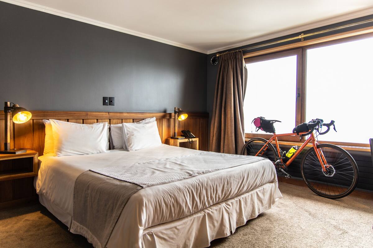 Une chambre d’hôtel avec un grand lit, une tête de lit en bois, deux tables de chevet avec des lampes, un vélo et une fenêtre avec des rideaux.