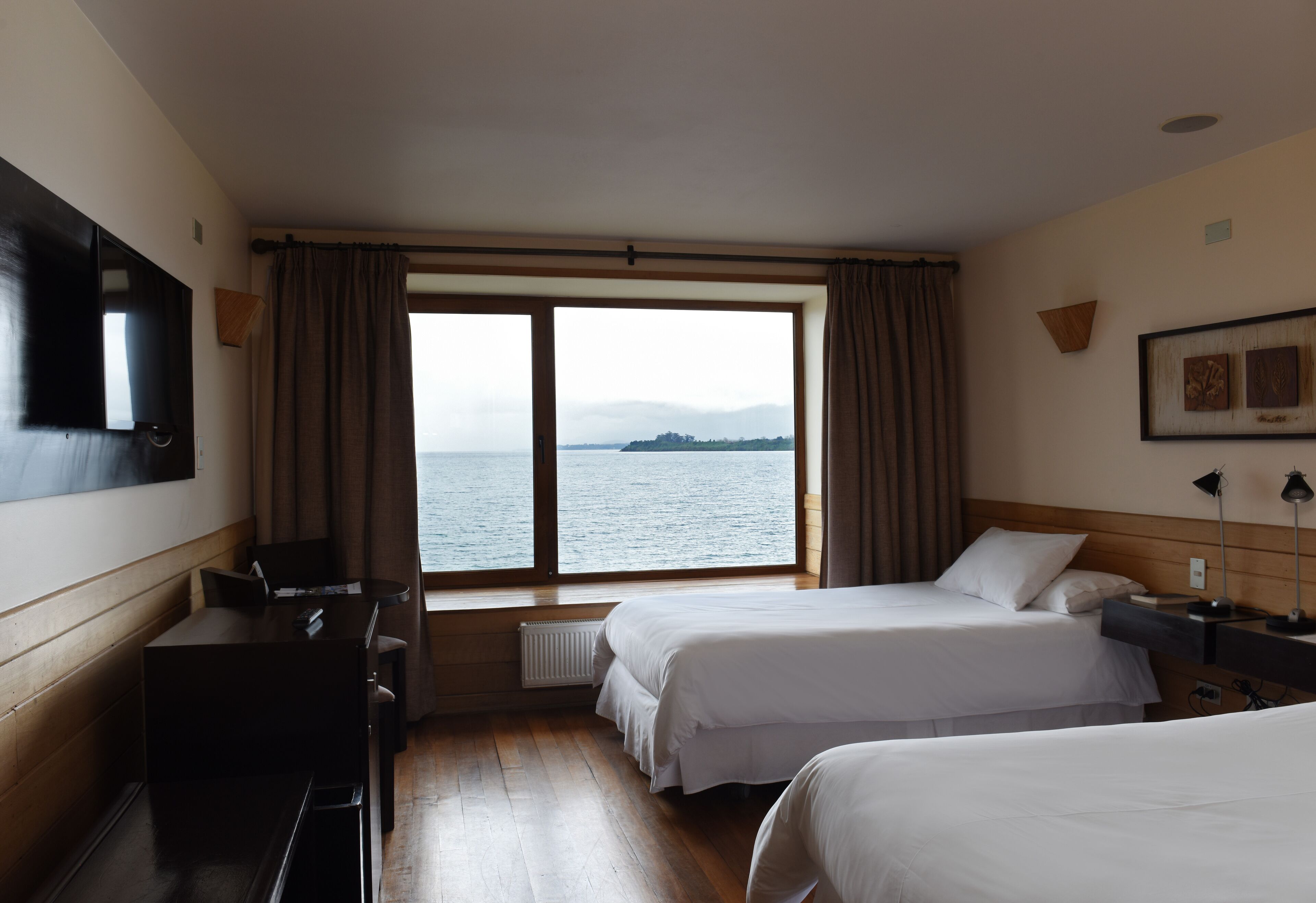 Een hotelkamer met twee bedden, een bureau, een stoel, een televisietoestel en een raam met uitzicht op zee.