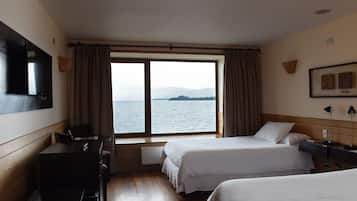 A hotel room with two beds, a desk, a chair, a television, and a window with a view of the sea.