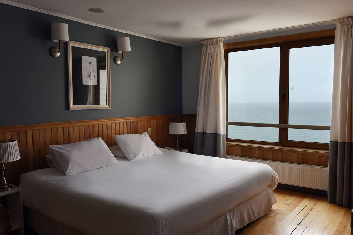 Une chambre d’hôtel avec un grand lit, une tête de lit en bois, deux lampes de chevet, une œuvre d’art encadrée, une fenêtre avec des rideaux et une vue sur la mer.