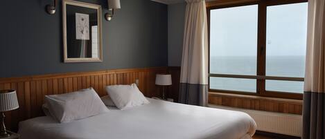 A hotel room with a large bed, wooden headboard, two bedside lamps, a framed artwork, a window with curtains, and a view of the sea.