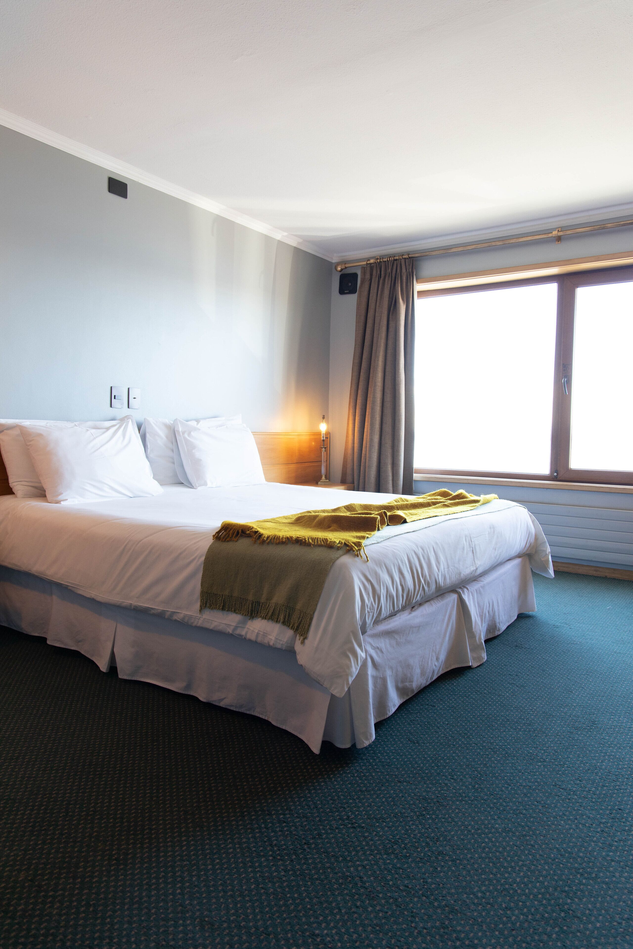 A hotel room with a large bed, a window with curtains, and a wall-mounted light.