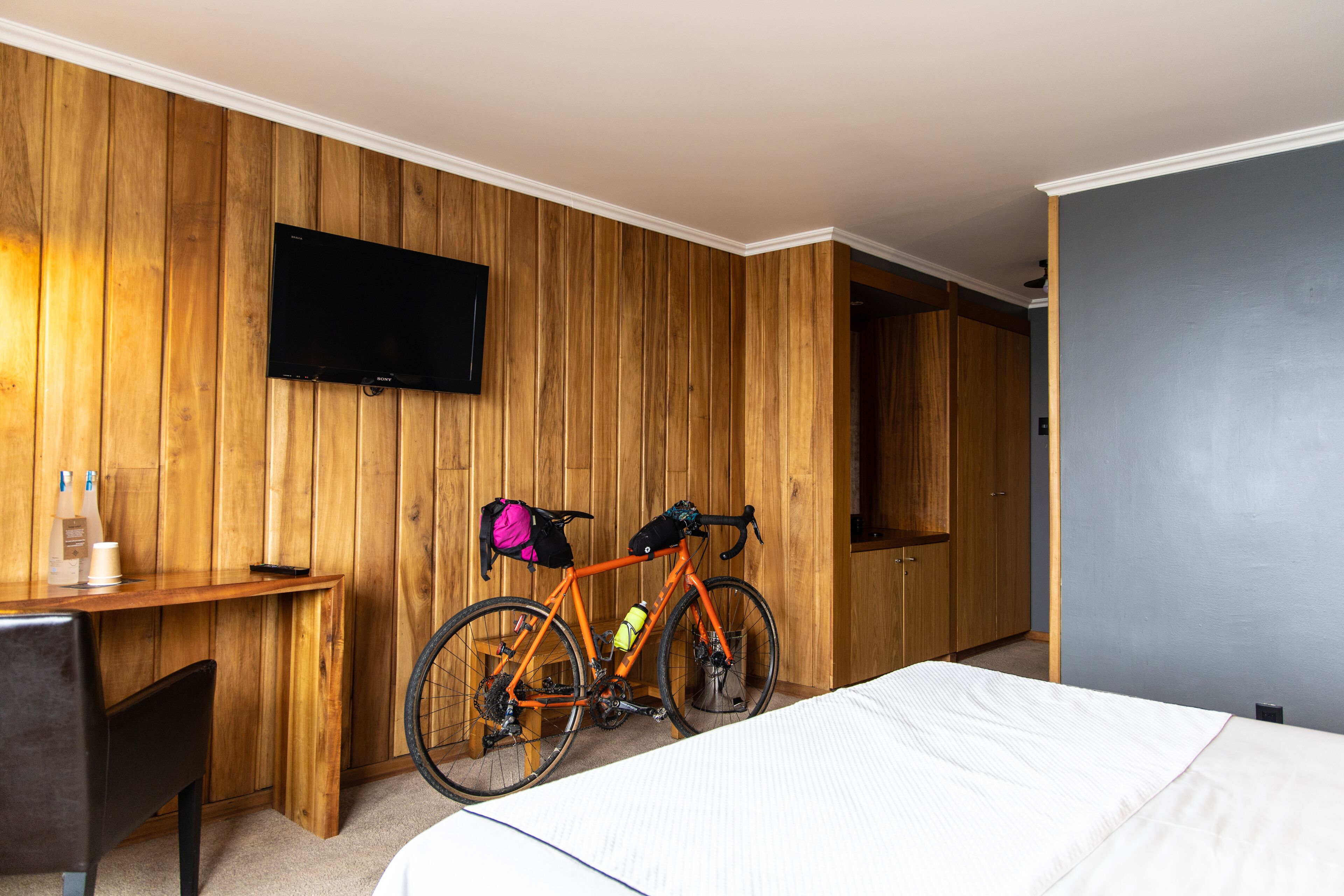 A hotel room with a wooden paneled wall, a mounted television, a bed, a desk with a chair, and a bicycle.
