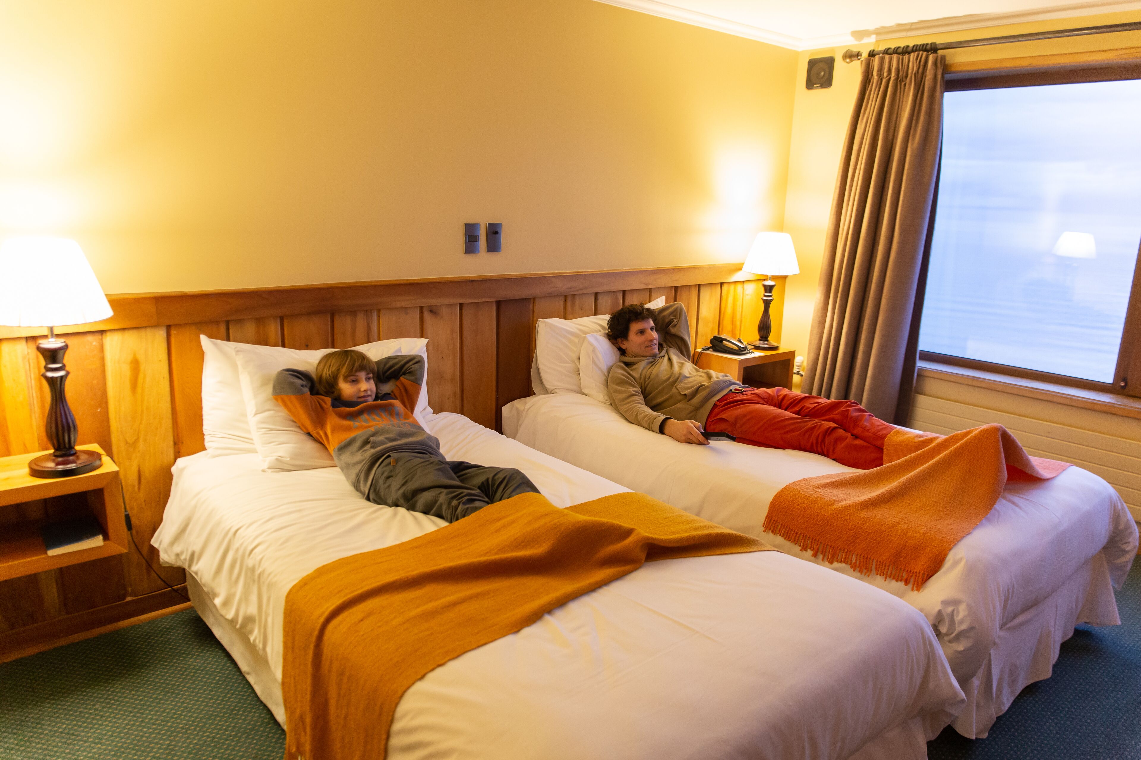 A hotel room with two beds, wooden headboard, bedside lamps, and a window with curtains.